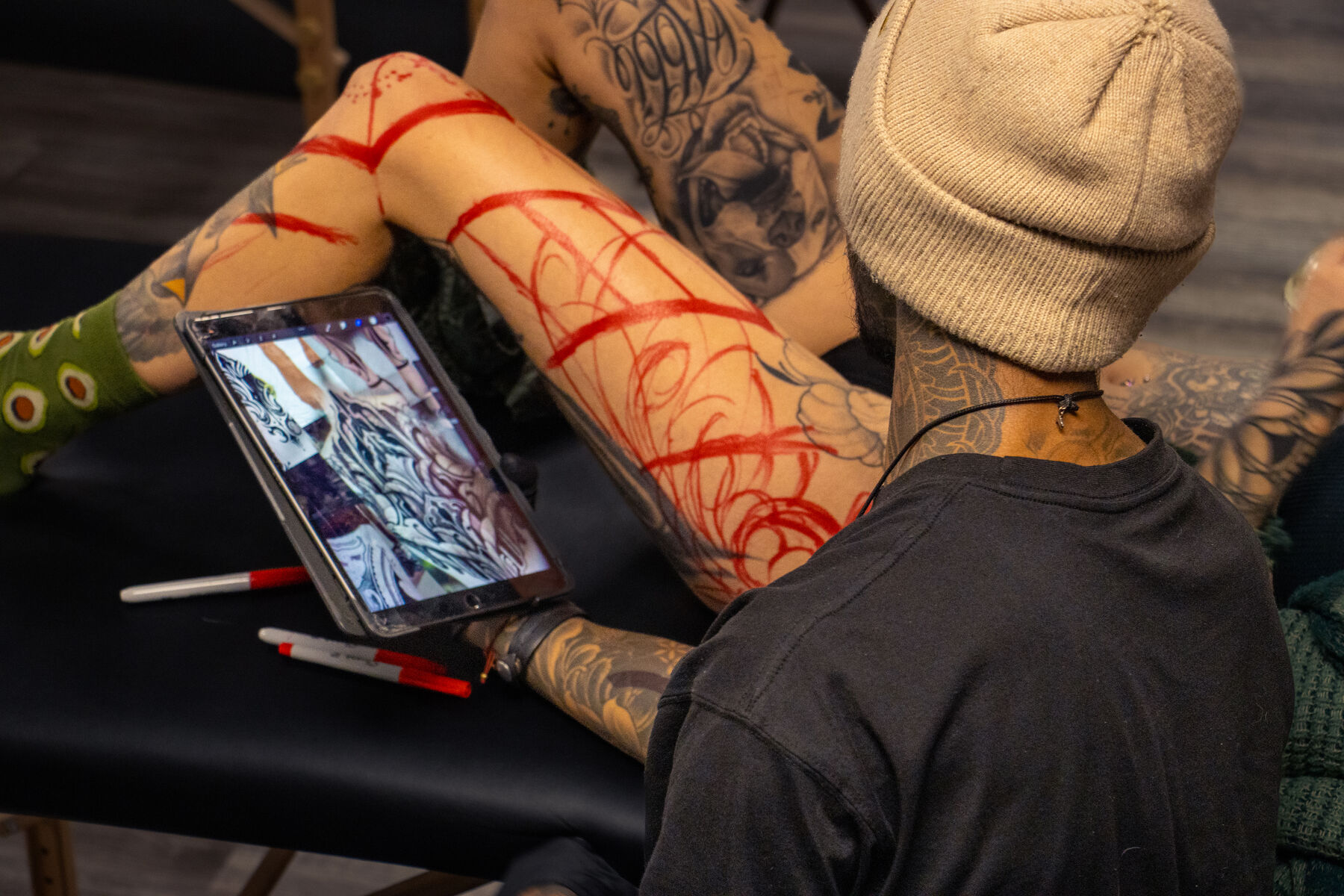 Artist checking a design on a tablet beside a freehand red stencil on a leg