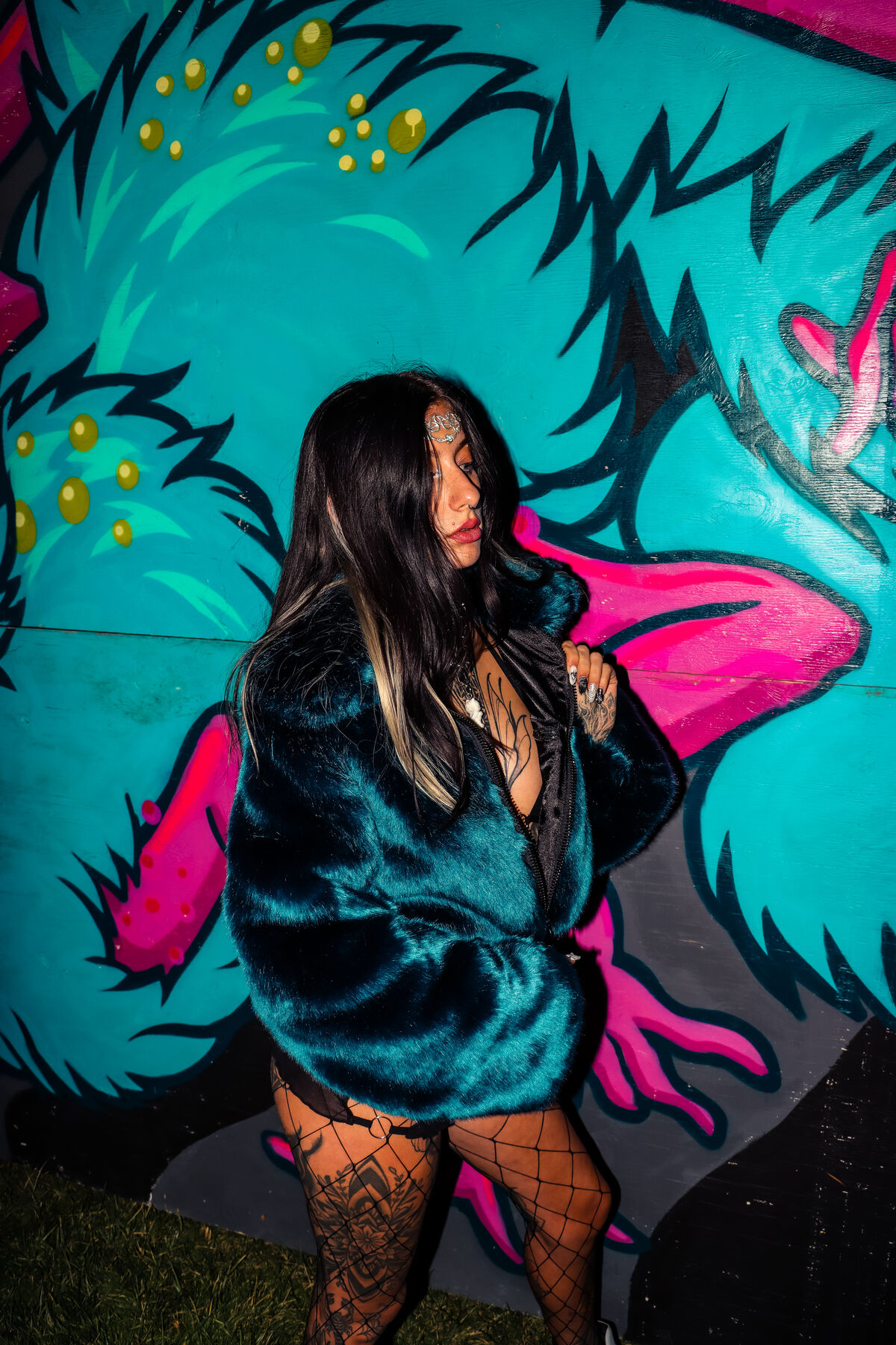 Ali in the dark green faux fur coat in front of a teal and pink mural