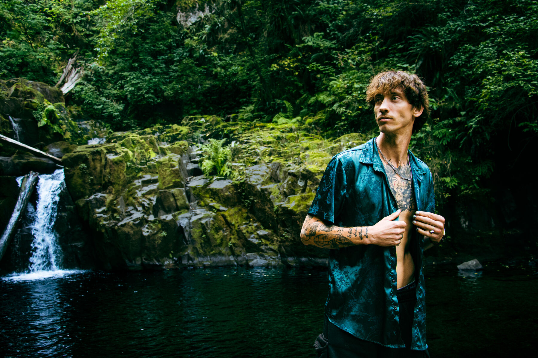 Brandon in a teal satin shirt standing in front of a small waterfall and mossy rock pool