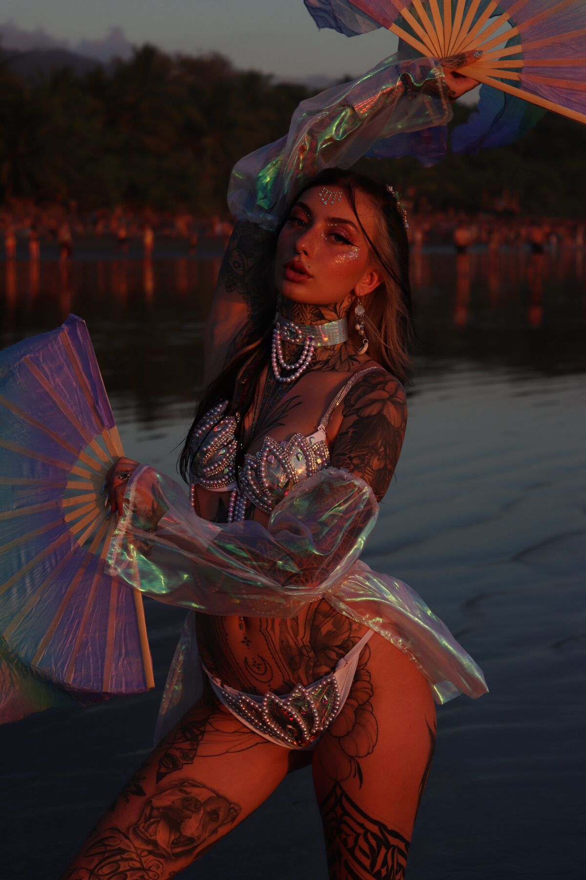 Ali holding an iridescent fan at the edge of a lagoon at dusk